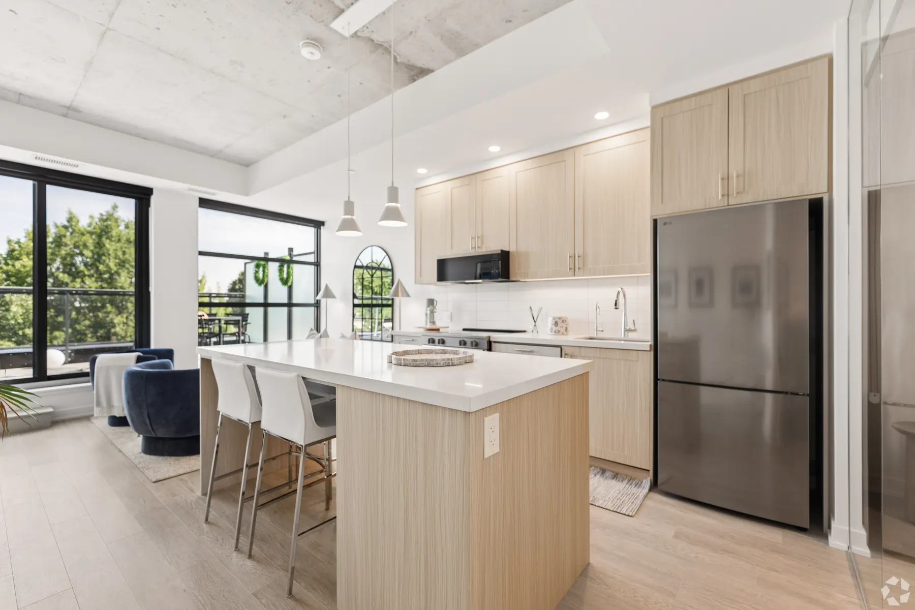 A one bedroom suite kitchen: pale oak cabinetry and a waterfall stone island with three stools under pendant lights, beneath an exposed concrete ceiling