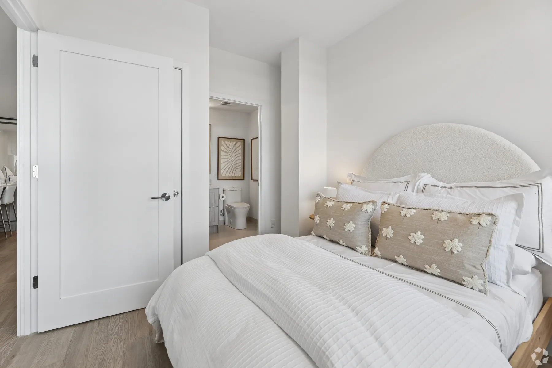 A suite bedroom: an arched upholstered headboard and white bedding against a white wall, with the ensuite through the open door