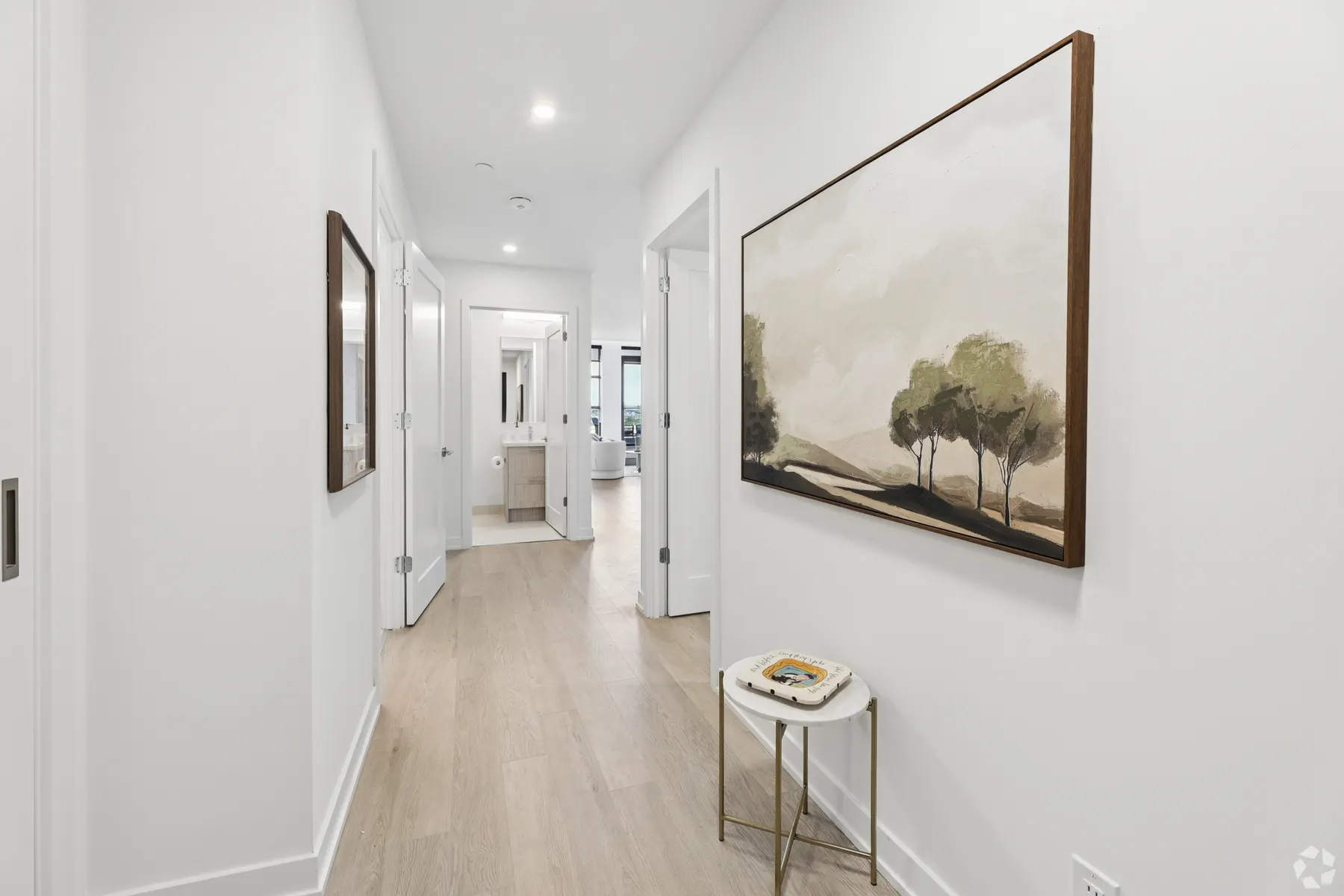 A suite hallway: a framed landscape above a small round table on pale plank flooring, bedroom and bathroom doors along the way