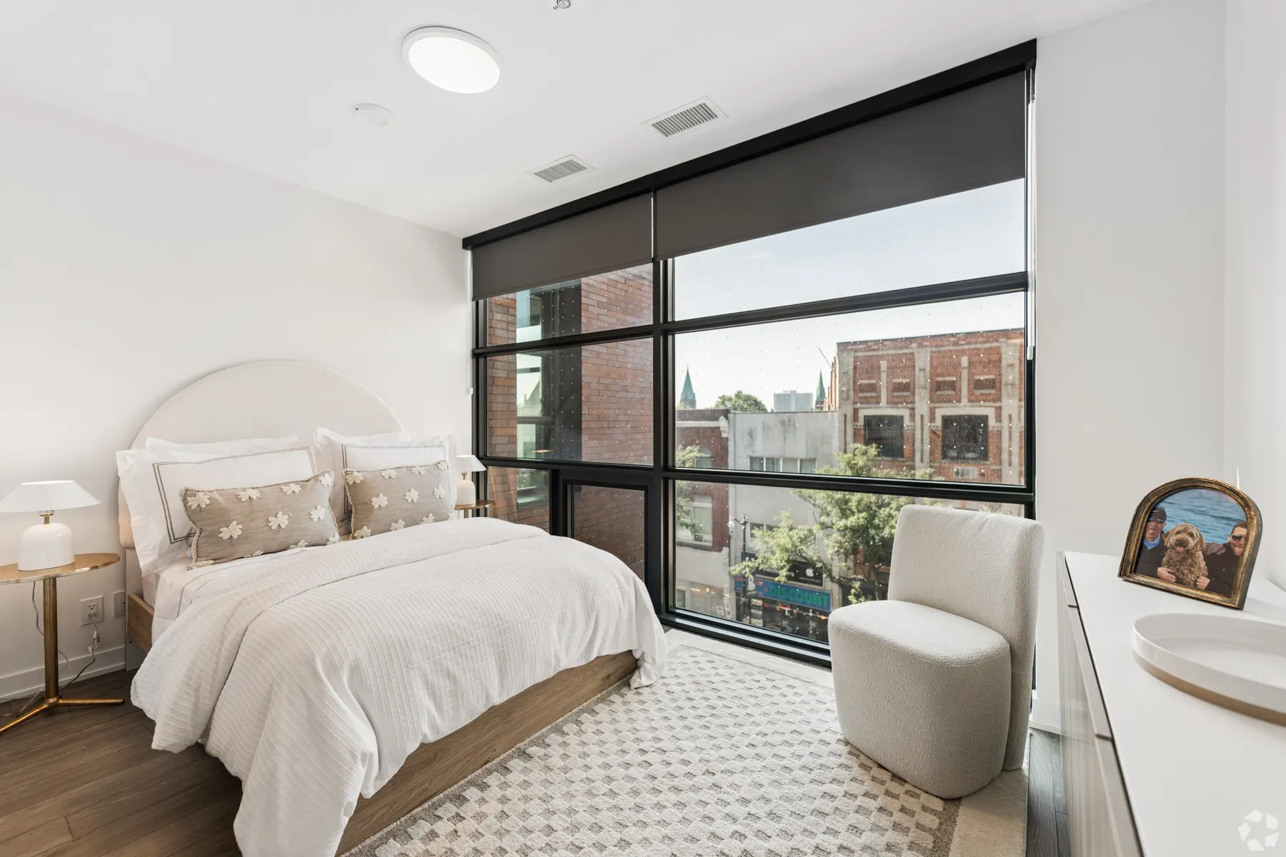 A two bedroom primary bedroom: an upholstered headboard against white walls beside a full height window onto rooftops and treetops