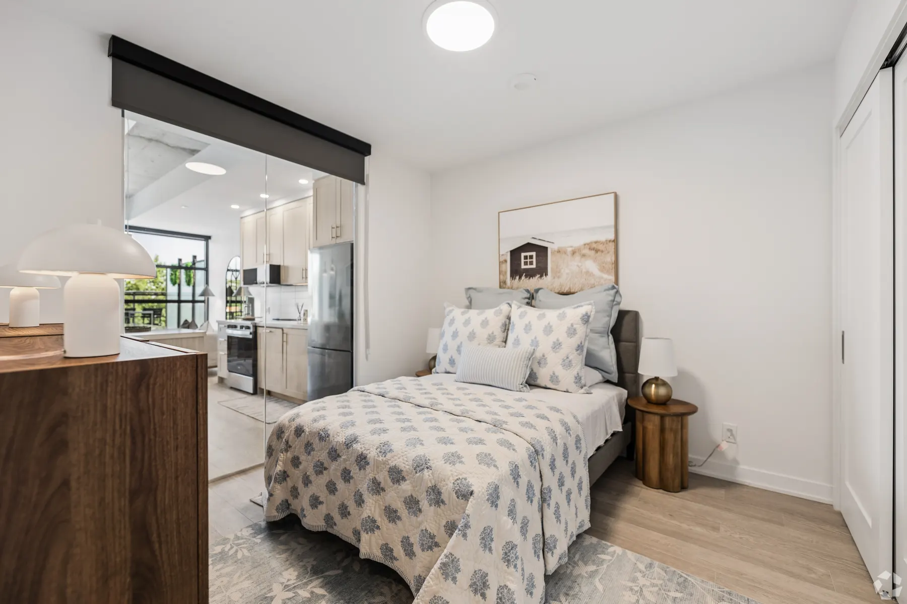 A one bedroom suite bedroom: a bed against a white wall beside a mirrored sliding wall reflecting the kitchen beyond