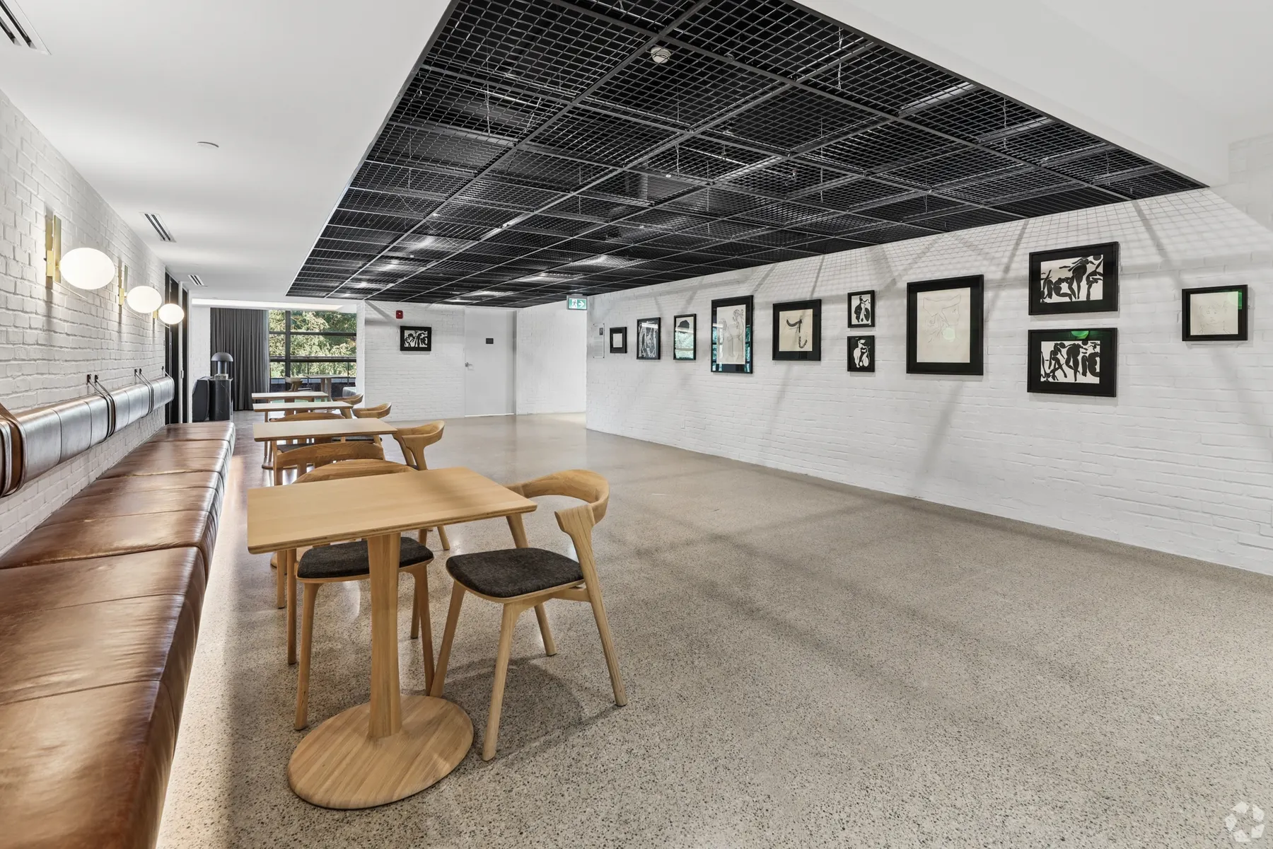 The café lounge along its length: oak tables and chairs beside a timber bench, a gallery wall of framed prints under the black grid ceiling
