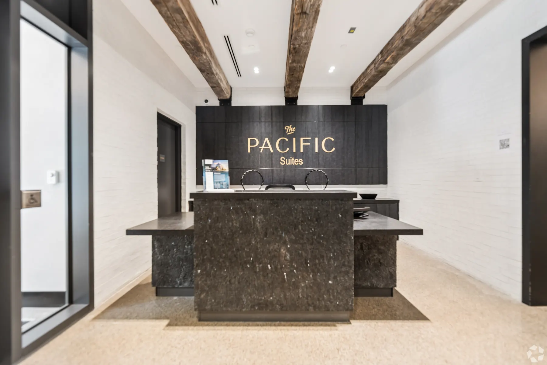 The concierge desk in dark veined stone beneath reclaimed timber beams, The Pacific Suites lettered in gold on the panelled wall behind