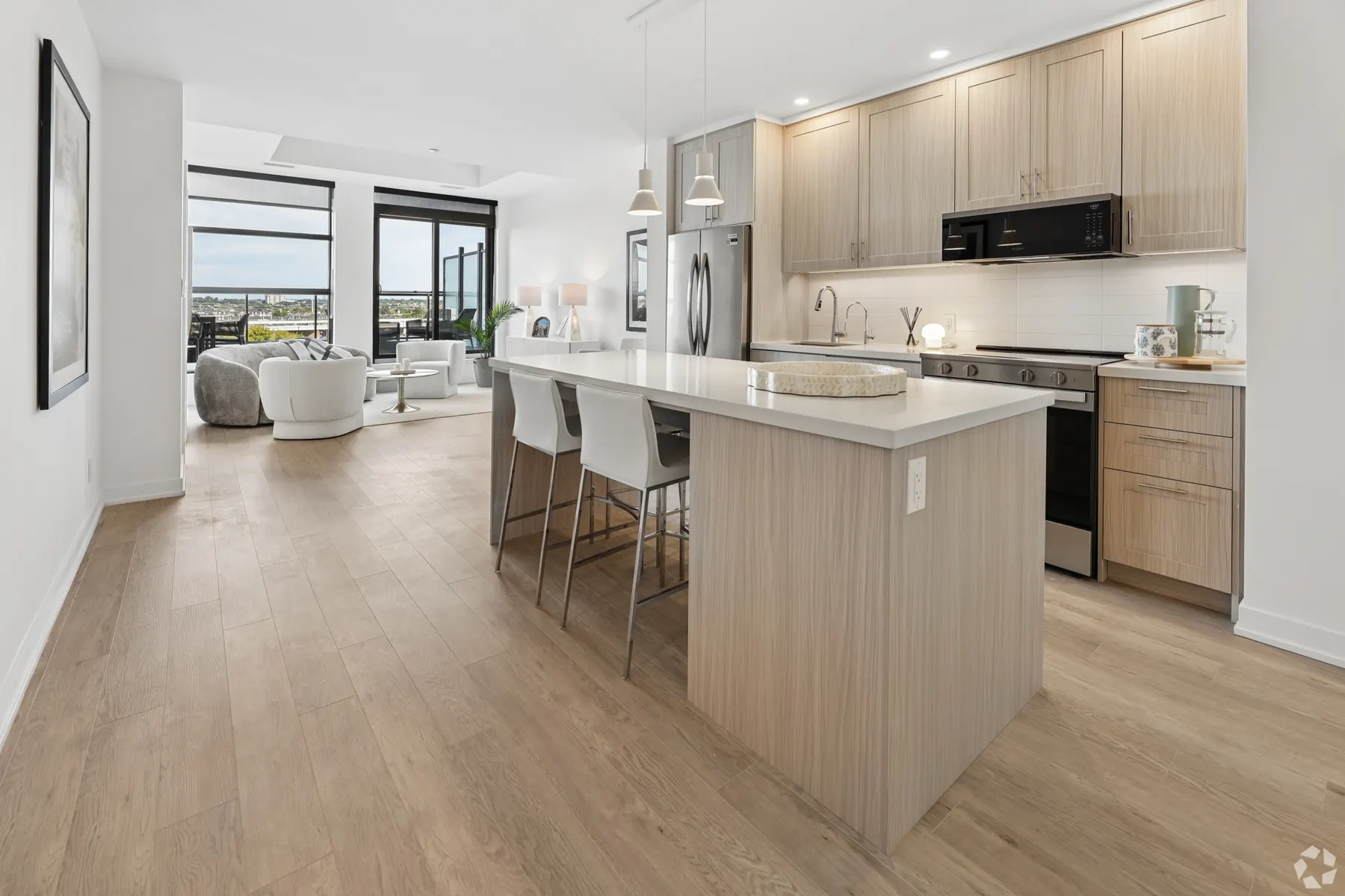 A suite kitchen and living room: a stone waterfall island with three stools under pendant lights, pale oak cabinetry and stainless appliances, the living room and its windows beyond