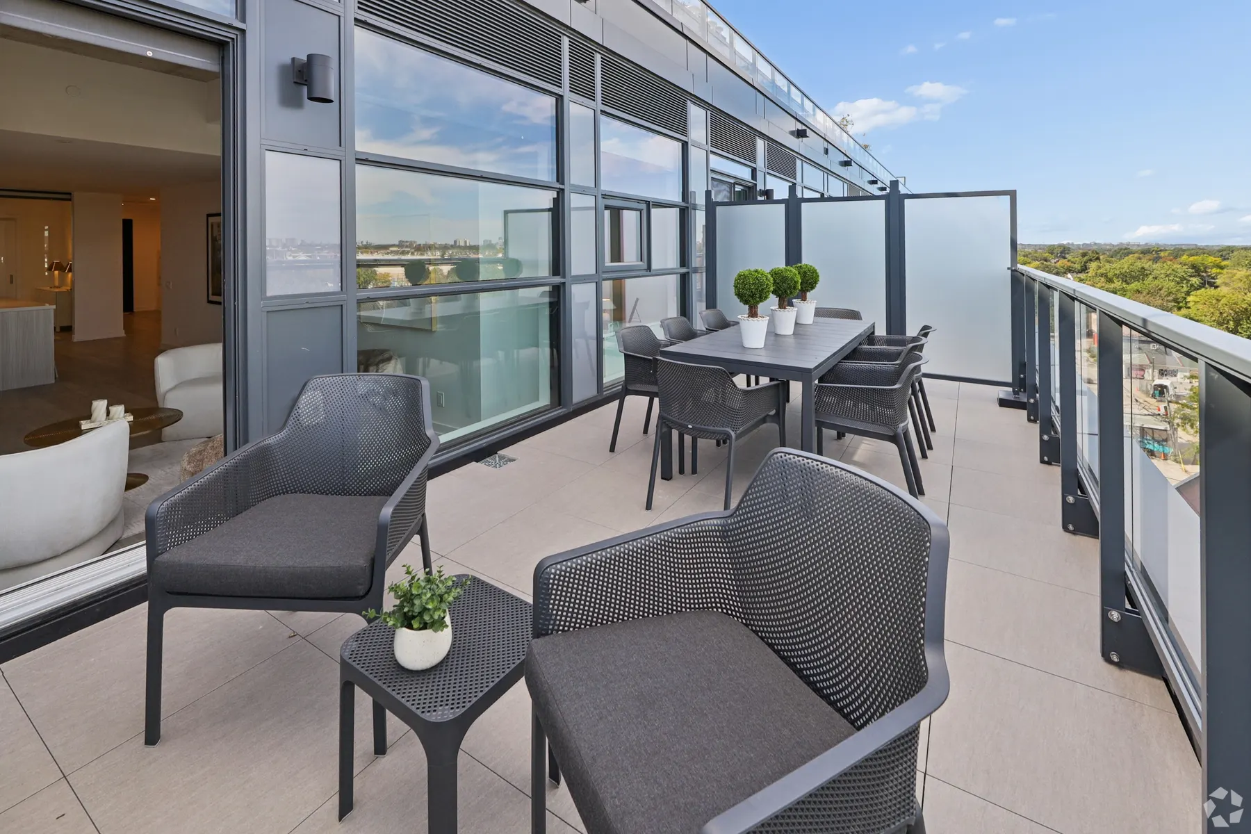 A suite terrace: woven armchairs and a dining table along a glazed balcony rail, the living room through the sliding door and the treetops beyond