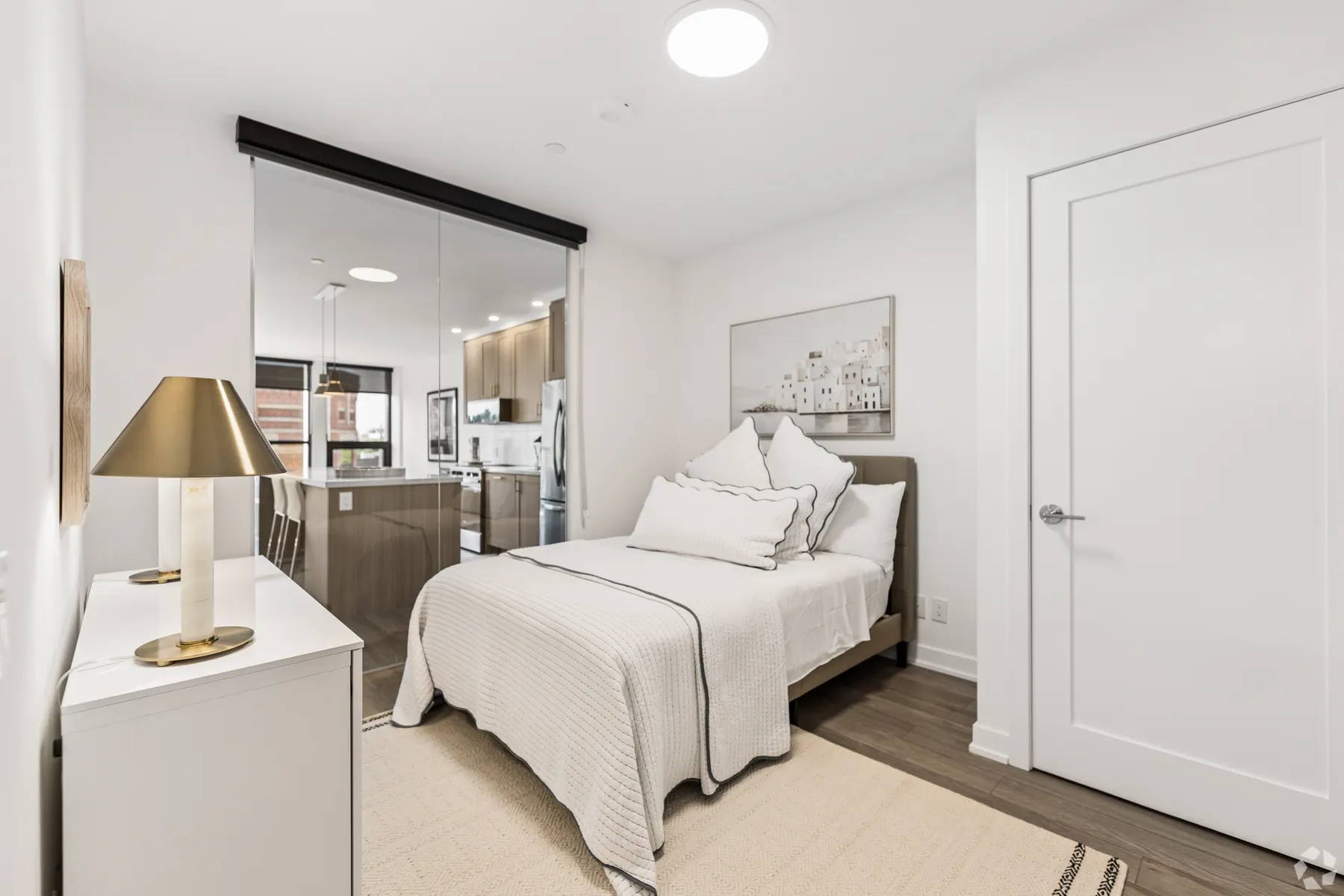 A second bedroom: a double bed against a white wall beside a mirrored sliding wall that reflects the kitchen, a white dresser and a brass lamp opposite