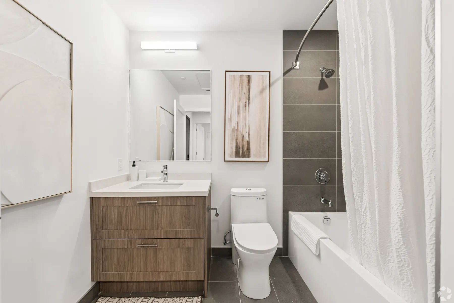 A second bathroom: a wood vanity with a white stone counter under a mirror, a tub behind a white curtain, tiled in grey