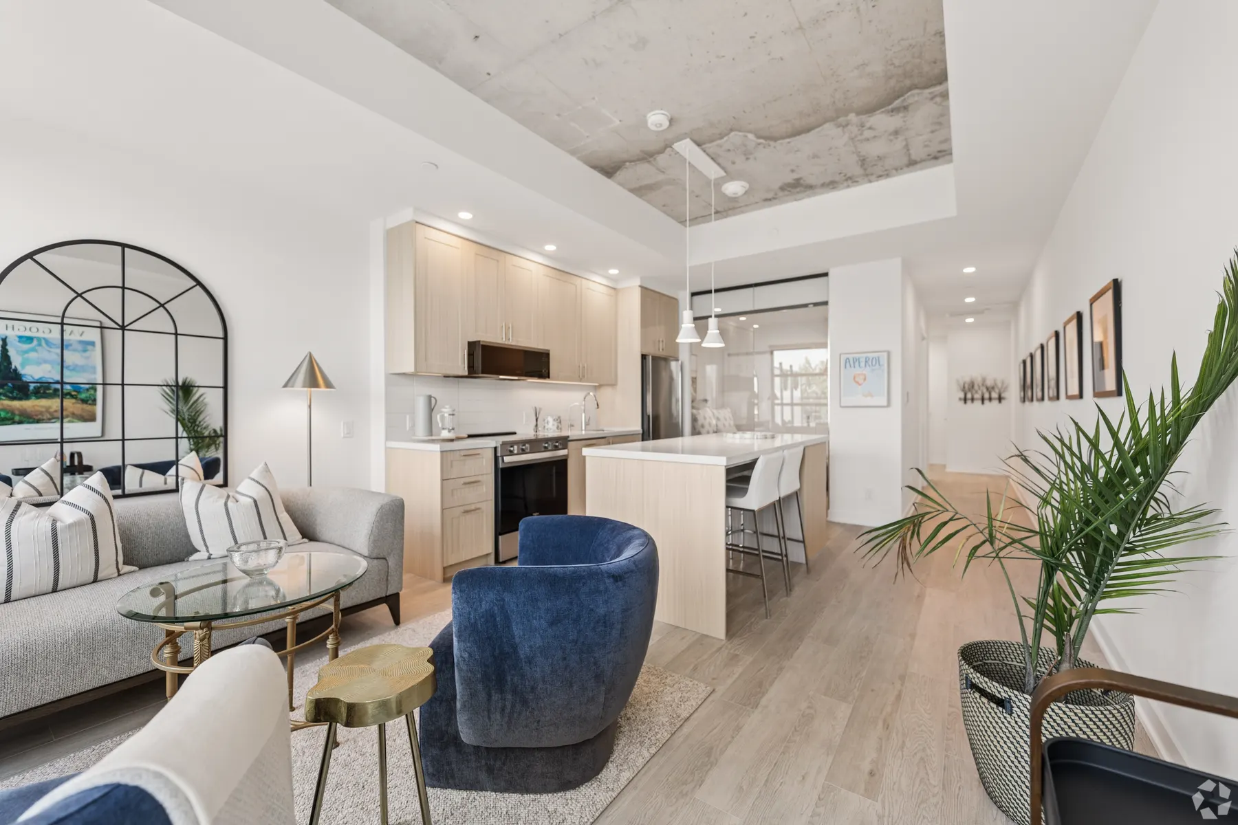 A one bedroom suite: an open living room with a grey sofa and a blue armchair under an exposed concrete ceiling, the pale oak kitchen beyond