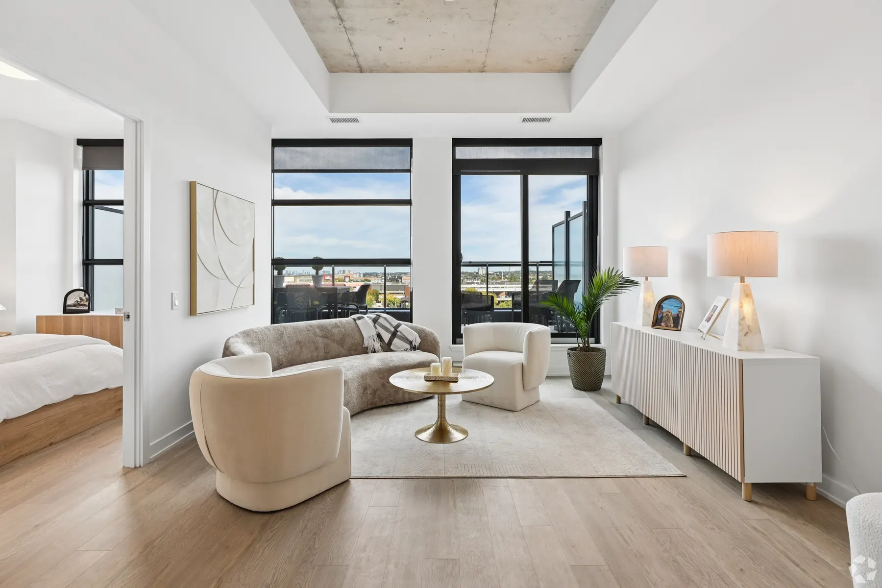 A suite living room: a curved bouclé sofa and two armchairs on a pale rug beneath a tray ceiling, tall windows onto rooftops and the bedroom through an opening to one side