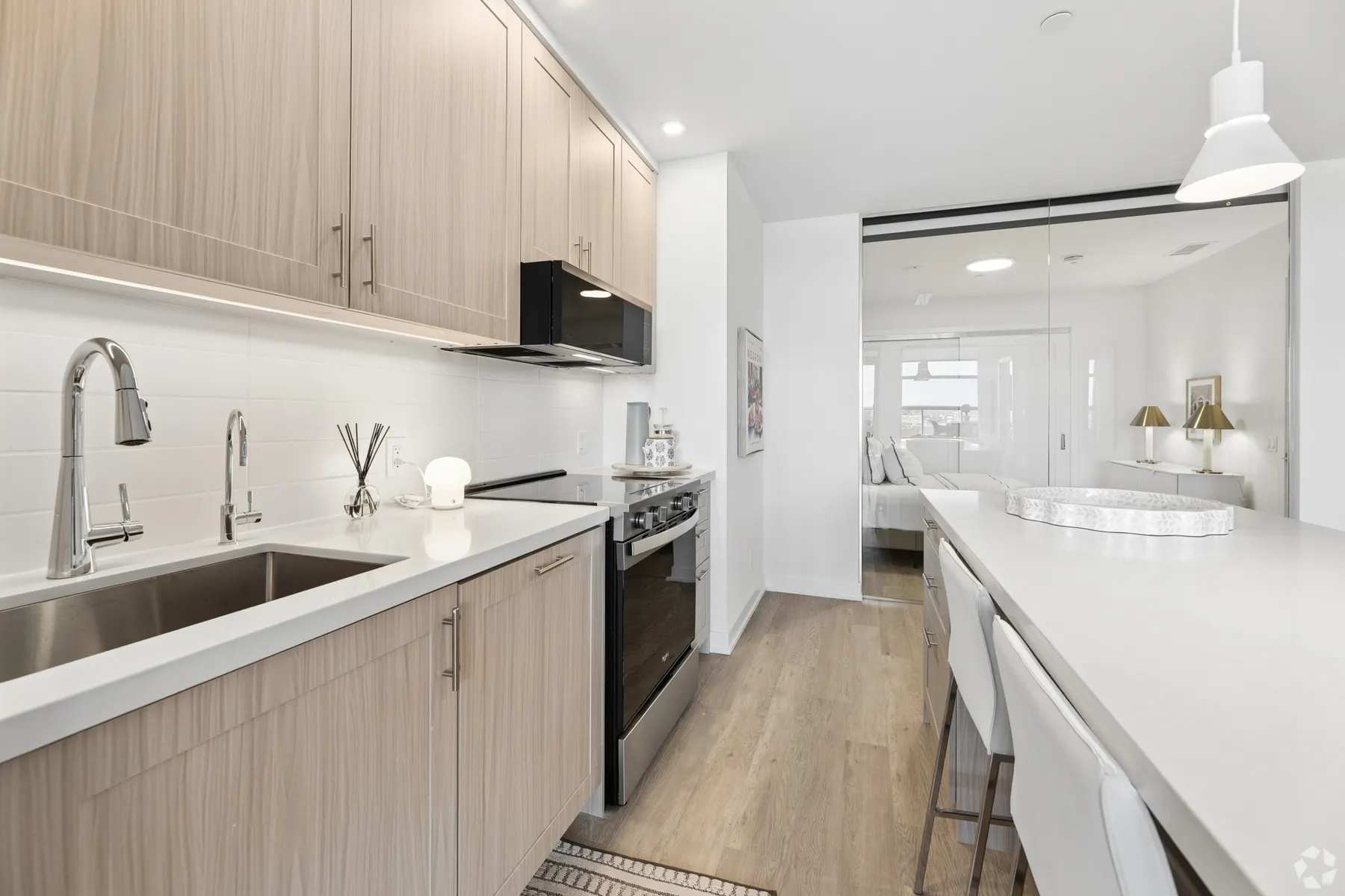 A suite kitchen: a stainless sink and a gas range under pale oak cabinetry, with the breakfast bar and the living room beyond