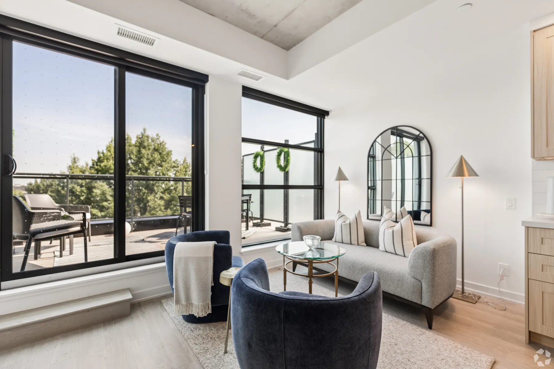 A suite living room opening onto a furnished balcony through two window walls, blue armchairs and a grey sofa around a glass table