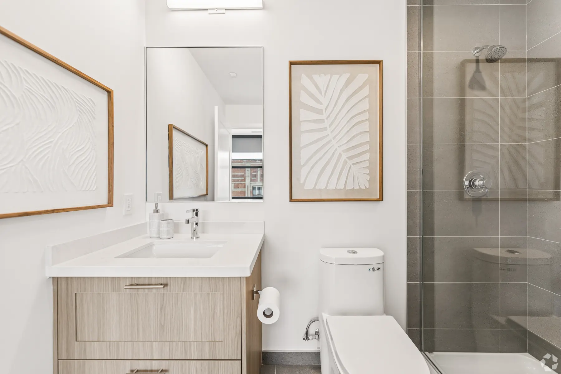 A primary ensuite: a white stone vanity under a framed mirror beside a glass walk-in shower, tiled floor to ceiling in grey