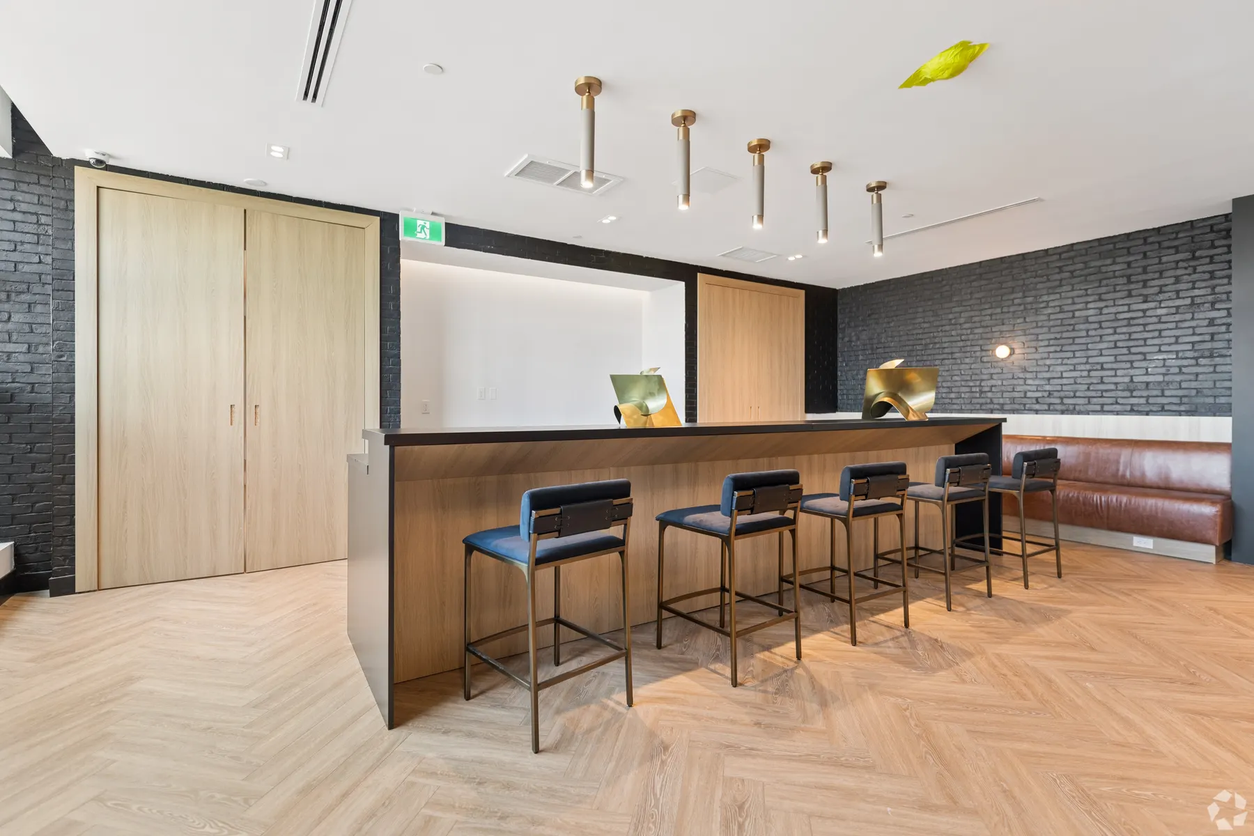 The lounge bar: a long counter with four stools under brass pendants, timber doors to one side and a leather banquette along the dark brick wall