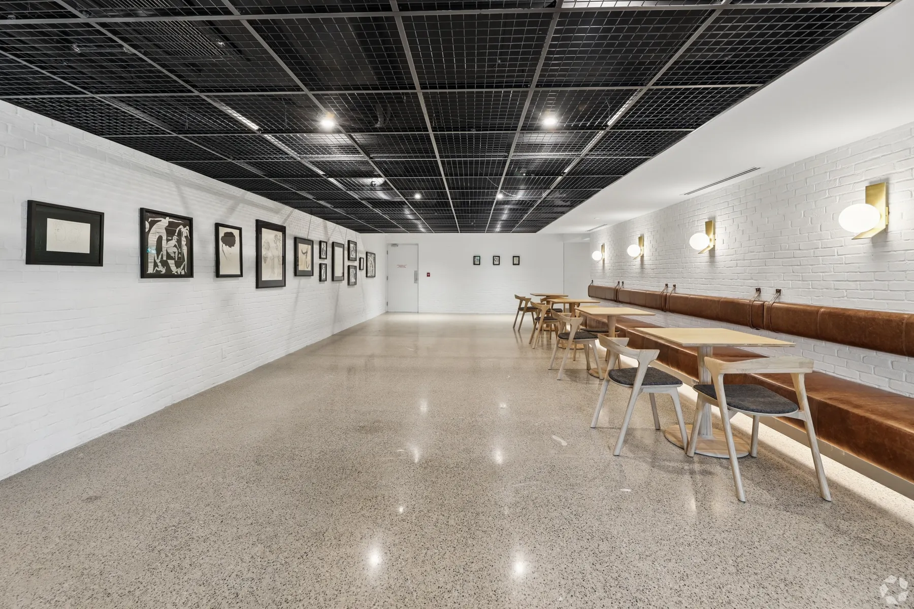 The café lounge from the entrance: long work tables and stools under a black grid ceiling, a timber bench along the white brick wall and a glazed meeting room at the far end